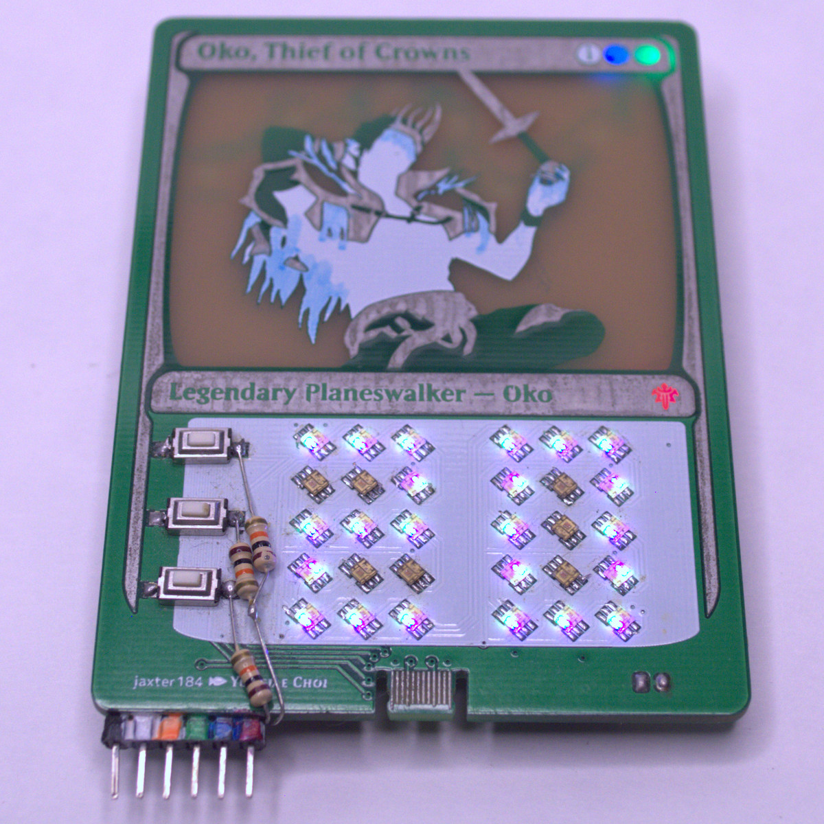 An older version on a green circuit board, with an image of the card etched into the PCB itself, prominently
	displaying the card art. The rules text area has two 3x5 matrices of LEDs displaying digits. There are also switches
	on the left of the LED matrices.