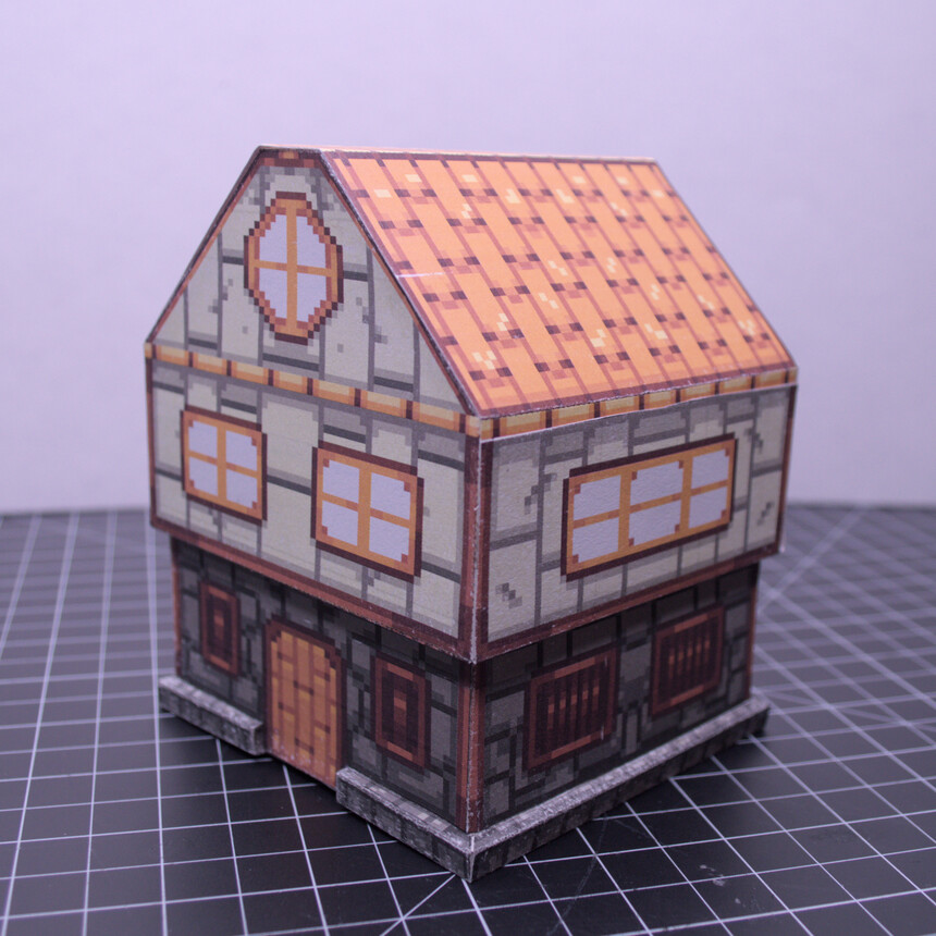 photo of a little papercraft house in a sort of Tudor Revival style, with a stone first floor, wooden door, and stucco second floor with wooden beams. very simple shapes, basically a cube with a triangular prism roof. first floor is very slightly inset from the rest of the house. It is textured in a pixel-art style