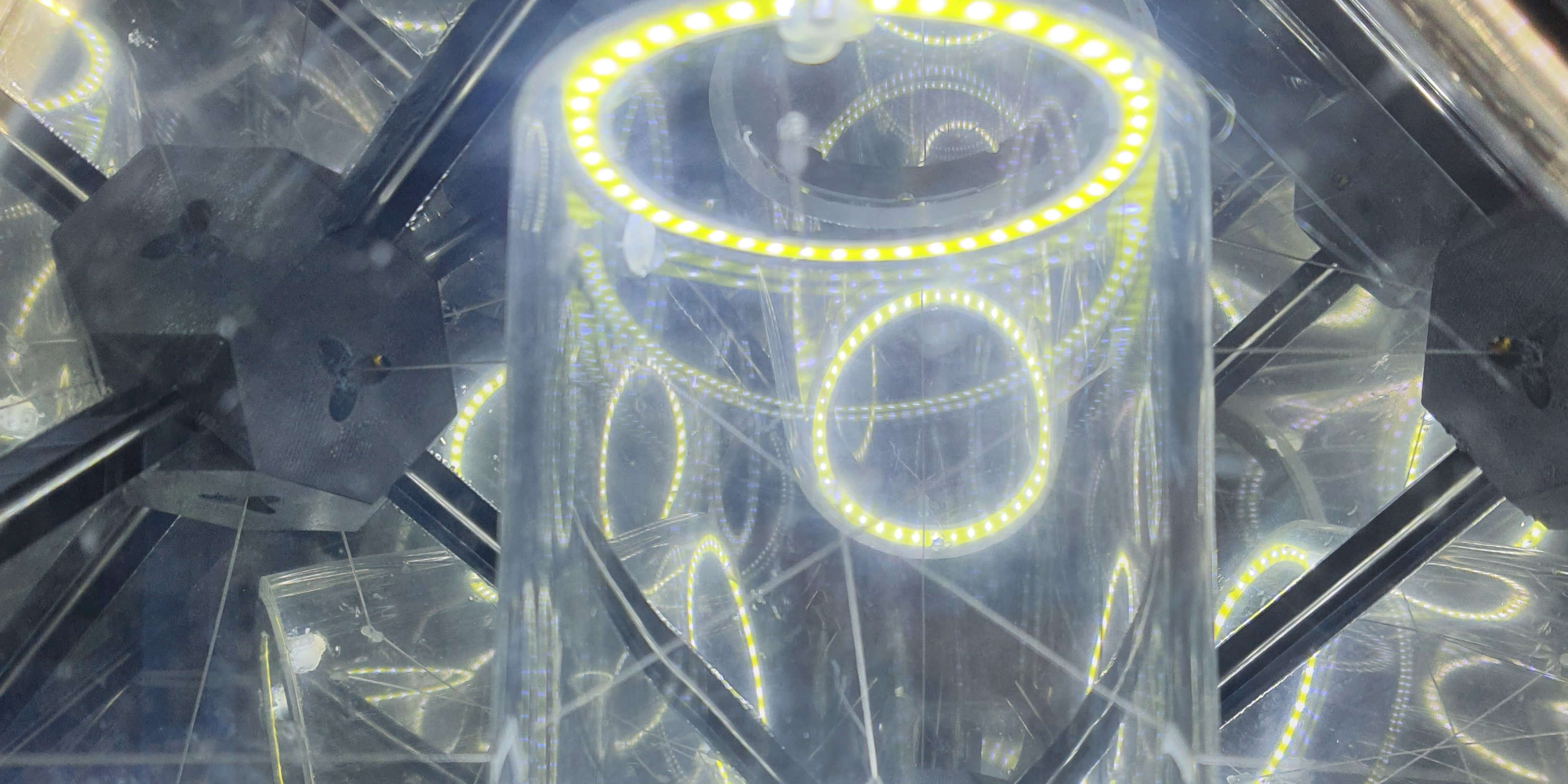 close-up image of a sculpture featuring an LED ring, being refracted by a clear tube and chaotically reflected off the inner walls of the cube enclosing it. Thin steel cables are faintly visible coming out of the black plastic corners of the cube.